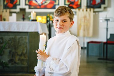 Küçük çocuk ilk kutsal komünyonunu alıyor. Vaftiz mumtutan mutlu çocuk. Katolik curch geleneği. Sunak yakınındaki bir kilisede beyaz geleneksel elbiseli çocuk