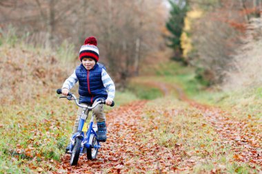 Sonbahar Ormanı Parkı 'nda renkli, sıcak giysiler içinde bisiklet süren küçük çocuk. Doğada güneşli bir sonbahar gününde bisiklet süren aktif bir çocuk. Güvenlik, spor, çocuk konseptiyle eğlence. Çocuklar için gündelik moda.
