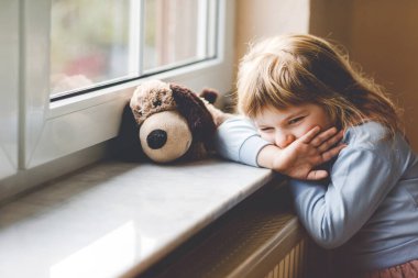 Üzgün küçük kız pencerenin kenarında oturup dışarı bakıyor. Üzgün ağlayan çocuk kendini mutsuz hissediyor. Corona virüsü salgını sırasında tecrit ve tecrit kavramı. Yalnız anaokulu çocuğu.