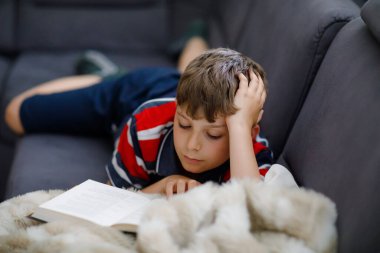 Şirin sarışın çocuk dergi ya da kitap okuyor. Heyecanlı çocuk sesli okuyor, kanepede oturuyor. Okul çocuğu, aile, eğitim. Covid zamanı evde eğitim.