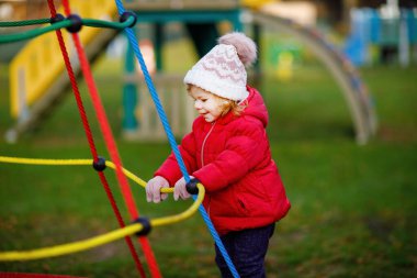 Küçük tatlı kız oyun alanında eğleniyor. Mutlu küçük çocuk tırmanışı, farklı ekipmanların üzerinde sallanmak ve kaymak. Renkli giysiler içinde soğuk bir günde. Çocuklar için aktif açık hava oyunu.