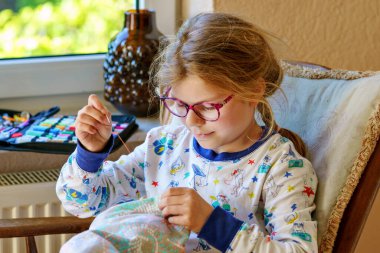 Küçük kız doldurulmuş hayvanı için yastık dikiyor. Mutlu Çocuk elinde iğne iplikle pencerenin yanında oturuyor..