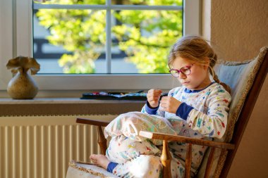 Küçük kız doldurulmuş hayvanı için yastık dikiyor. Mutlu Çocuk elinde iğne iplikle pencerenin yanında oturuyor..