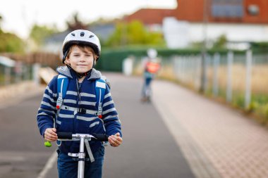 Kasklı aktif okul çocuğu güneşli bir günde şehirde scooterıyla geziyor. Renkli elbiseli mutlu çocuk okula giderken bisiklete biniyor. Çocuklar için güvenli okul yolu..