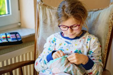 Küçük kız doldurulmuş hayvanı için yastık dikiyor. Mutlu Çocuk elinde iğne iplikle pencerenin yanında oturuyor..