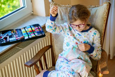 Küçük kız doldurulmuş hayvanı için yastık dikiyor. Mutlu Çocuk elinde iğne iplikle pencerenin yanında oturuyor..