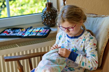 Küçük kız doldurulmuş hayvanı için yastık dikiyor. Mutlu Çocuk elinde iğne iplikle pencerenin yanında oturuyor..