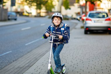 Kasklı aktif okul çocuğu güneşli bir günde şehirde scooterıyla geziyor. Renkli elbiseli mutlu çocuk okula giderken bisiklete biniyor. Çocuklar için güvenli okul yolu..