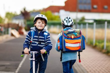 Güneşli bir günde, ellerinde scooterla şehirde gezen kasklı iki okul çocuğu. Renkli giysiler içinde okula giderken bisiklet süren mutlu çocuklar. Çocuklar için güvenli okul yolu..