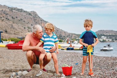 Mutlu büyükbaba ve iki küçük çocuk kumsalda oynuyorlar ve kumdan ve taştan kale yapıyorlar. Güzel bir aile deniz ve okyanusta tatilin tadını çıkarıyor. Büyükbaba ve çocuklar.