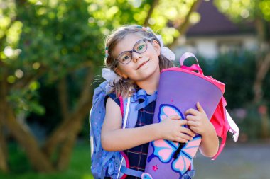 Gözlüklü ve mavi bir sırt çantası olan, denizci üniforması giyen küçük bir kız, Schultuete adında renkli bir okul konisi ve sırt çantası taşıyor. Mutlu çocuk gülümsüyor, okulun ilk günü için hazır.