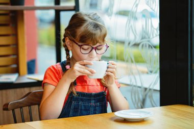 Gözlüklü, hoş bir kafede kahvaltı sırasında sıcak çikolata içen sevimli küçük kız. Mutlu çocuk sıcak içkinin tadını içeride çıkarıyor. Gerçek hayat, gerçek insanlar çocukluk rahatlığı anı