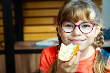 Gözlüklü sevimli ilkokul kızı kahvaltıda tereyağlı ekmek yiyor. Sabah yemeğinin tadını çıkaran mutlu çocuğun yakın plan portresi. Gerçek hayat, gerçek insanlar çocukluk sevinci anı.