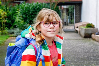 Gözlüklü ve sırt çantalı şirin bir ilkokul öğrencisi okula giderken yolda yürüyor. Gülümseyen öğrenci okula, eğitime ve çocukluk yaşam tarzı konseptine hazır..