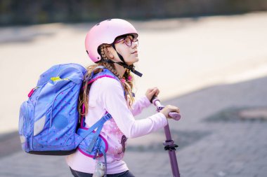 Okula giderken scooter 'a binen gülümseyen küçük okul kızı. Çocuk dışarıda kask ve sırt çantası takıyor. Okula güvenli ve mutlu bir rota, gerçek yaşam tarzı anı..