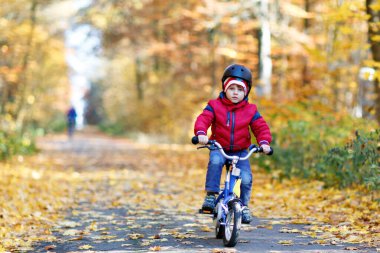 Sonbahar Ormanı Parkı 'nda renkli, sıcak giysiler içinde bisiklet süren küçük çocuk. Doğada güneşli bir sonbahar gününde bisiklet süren aktif bir çocuk. Güvenlik, spor, çocuk konseptiyle eğlence. Çocuklar için gündelik moda.