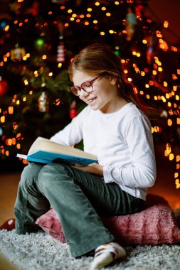 Küçük liseli kız yılbaşı ağacının yanında ışıklarla kitap okuyor. Mutlu çocuk, aile tatili. Okul çocuğu kitap okuyor.