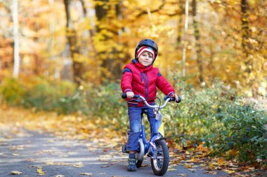 Sonbahar Ormanı Parkı 'nda renkli, sıcak giysiler içinde bisiklet süren küçük çocuk. Doğada güneşli bir sonbahar gününde bisiklet süren aktif bir çocuk. Güvenlik, spor, çocuk konseptiyle eğlence. Çocuklar için gündelik moda.