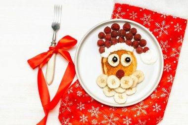 Christmas Santa Claus shaped pancake with sweet fresh raspberry berry and banana on plate, red sock, fork on white wooden background for kids children breakfast. xmas food with new year decorations.