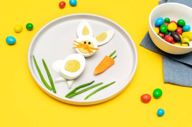 Easter Funny Creative Healthy breakfast lunch food idea for kids children. Bunny rabbit made from boiled eggs,peeled carrots, greens on plate yellow table background. Top view Flat lay.
