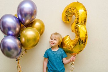 Blond cute happy Child kid boy celebrating third birthday with colorful balloons at party,home in front of wall.baby smiling looking at camera.adorable caucasian baby.Festive background decoration.