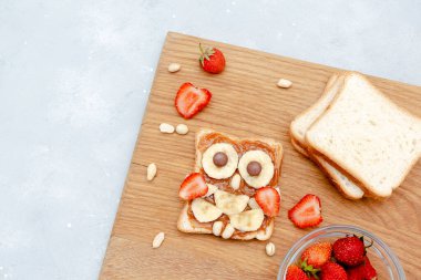 Komik baykuş suratlı tost ekmeği, fıstık ezmesi, muz, ahşap tahtada çilek. Tatlı çocuklar bebeğin tatlı tatlısını, sağlıklı kahvaltısını, öğle yemeğini, gri arka planda yemek sanatını, üst manzarayı....