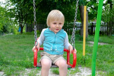 Cute funny blond little young toddler kid child boy sitting and riding on swing in playground in park.Children physical,emotional development and childhood daycare, kindergarten concept.