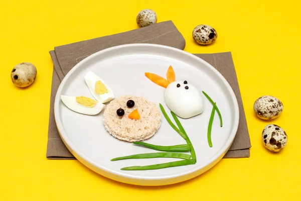 Easter funny creative healthy breakfast lunch food idea for kids, children.Bunny, rabbit made from boiled chicken eggs,bread, peeled carrots, greens on plate yellow table background.Top view Flat lay.