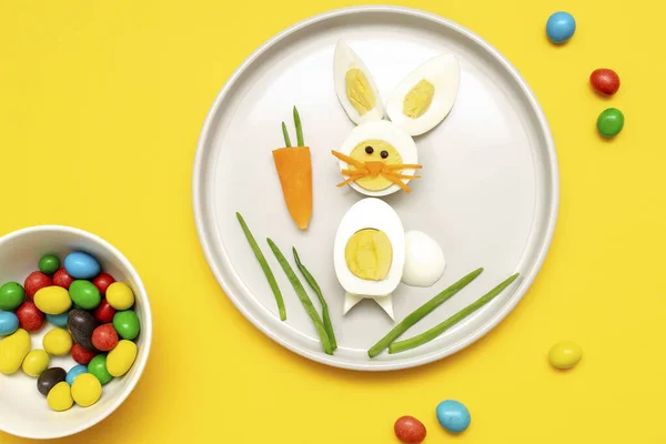 Easter funny creative healthy breakfast lunch food idea for kids, children.Bunny, rabbit made from boiled chicken eggs,peeled carrots, greens on plate yellow table background.Top view Flat lay.