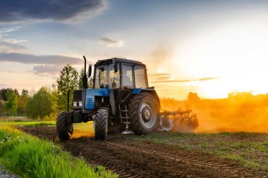 Günbatımında ilkbaharda çiftlikte tarım tarlası sürerken modern mavi traktör makineleri. Çiftçilik, bitki, mahsul, kırsal kesimde doğa ekmeden önce toprak ekimi yapmak..