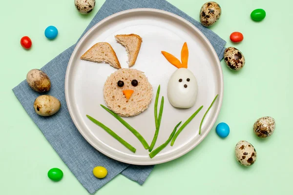 Easter funny creative healthy breakfast lunch food idea for kids, children. Bunny, rabbit made from boiled chicken eggs,bread, peeled carrots, greens on plate green table background.Top view Flat lay.