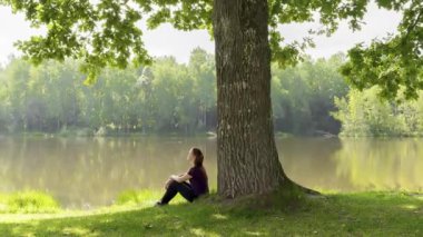 Teen young girl woman sitting on grass under huge high big old oak,trunk tree near lake,river, meditating,relaxing and smiling in National Park.inspiration nature beauty harmony landscape outdoors.