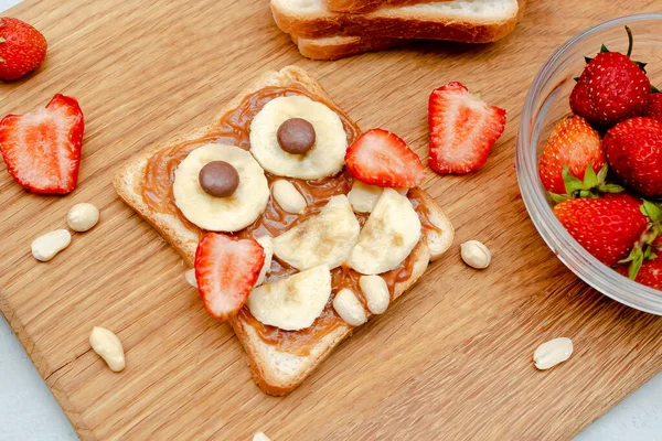 Komik baykuş suratlı tost ekmeği, fıstık ezmesi, muz, ahşap tahtada çilek. Tatlı çocuklar bebeğin tatlı tatlısını, sağlıklı kahvaltısını, öğle yemeğini, gri arka planda yemek sanatını, üst manzarayı....