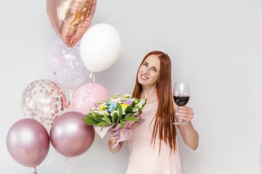 Mutlu, genç, çekici, kızıl saçlı kadın elinde balonlar, çiçek buketi, şarap kadehi ev partisinde doğum gününü kutlarken gülümsüyor..