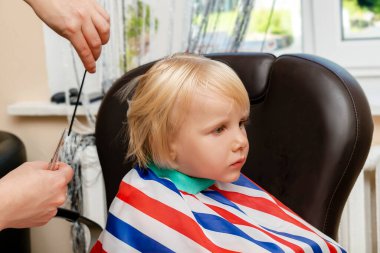 Little blond child boy getting haircut in beauty salon,barbershop. Hairdresser stylist cutting hair with professional tools-comb and scissors.Kids spa.Style and fashion,cutting hair for children.