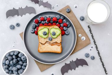 Funny scary monster face smile on halloween sandwich toast bread with peanut butter, blueberry, raspberry on plate,bats on gray background close up. Kids child sweet dessert breakfast lunch food.