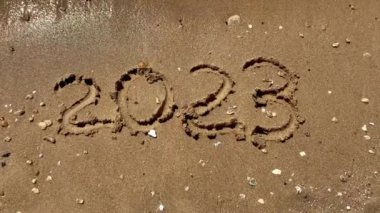 Metin rakamları 2023. Turistler tarafından altın kumlu sahil yüzeyi, deniz kıyısı, köpüklü deniz dalgalarıyla silinmiş kelimeler. Yeni yıl 2024 geliyor, kararlar, geri sayım..
