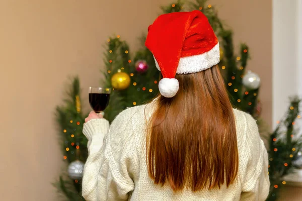 Happy woman standing against Christmas tree at home with wine glass.red hair lady in santa claus hat celebrating new year.Merry Christmas and Happy Holidays.