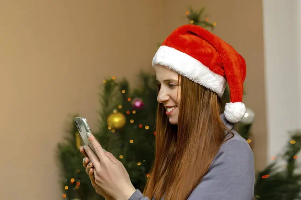 Happy beautiful woman against Christmas tree at home.Cheerful positive lady in santa claus hat using smartphone app call virtual chat online video Webcam.Merry Christmas,Happy Holidays Celebration.