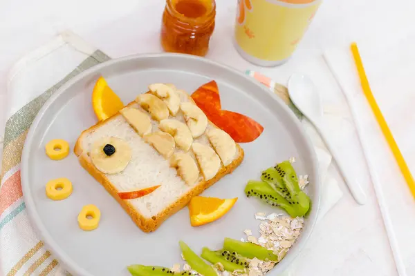 stock image Funny cute fish shape sandwich toast bread with banana, apple,orange, milk,honey. Kids childrens baby's sweet dessert healthy breakfast lunch food art on plate,wooden background close up.