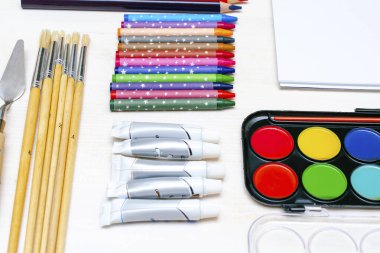 Suluboya boyaları, boya fırçaları, renkli kalemler, pastel boya kalemleri, boş suluboya kağıt pedler. Yaratıcılık yaratma süreci. Sanatçılar beyaz masaya bir şeyler çizerler. Malzemelerin üst görünümü..