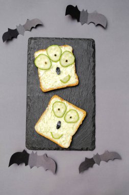 Cadılar bayramında kızarmış ekmek, yeşil avokado peynirli ricotta yağı, salatalıklar servis panosunda, yarasalar gri zeminde. Çocuklar için öğle yemeği..