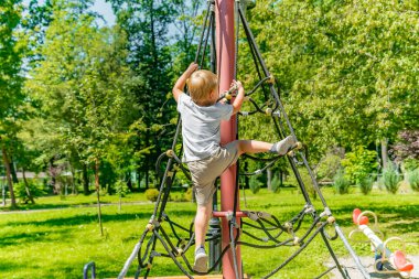 Küçük sevimli çocuk modern oyun bahçesinde ip merdivene tırmanıyor. Açık havada çocuklar için sokak sporu aktiviteleri, fiziksel gelişim, çocukluk, anaokulu..