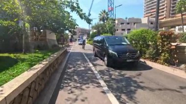 Bisikletçiler ve minibüs güneşli bir Monte Carlo caddesinde yol alıyorlar.