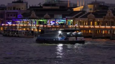 Tekne gece aydınlık nehir kenarındaki binaların önünden geçiyor.