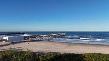 İnsansız hava aracı görüntüsü, berrak gökyüzünün altında sakin bir Altın Sahil sahili ve iskeleyi yakalıyor. Geniş okyanusu ve kıyı manzarasını vurguluyor.