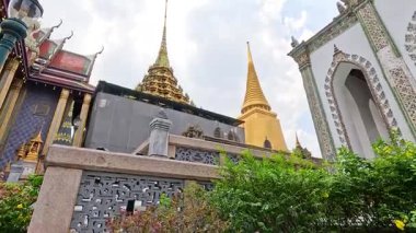 Wat Phra Kaew 'in Bangkok' taki altın kulelerinin dinamik görüntüsü, parlak bir gökyüzü altında karmaşık bir mimari sergiliyor.