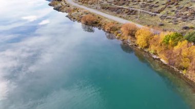 Drone, Dunstan Gölü, Cromwell, Yeni Zelanda boyunca dingin sonbahar manzarasını canlı yapraklarla yakalar. Sakin su gökyüzünü ve ağaçları yansıtır.