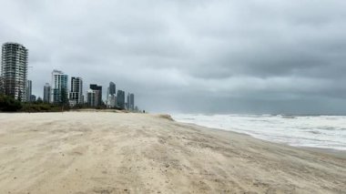 Şiddetli rüzgarlar ve çalkantılı dalgalarla dolu fırtınalı bir sahil sahnesi bulutlu gökyüzünün altındaki şehir binalarının arka planına kurulmuş.