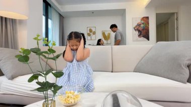 Modern bir oturma odasında ebeveynler tartışırken genç bir kız koltukta kulaklarını kapatıyor. Parlak ışıklandırma gergin atmosferi güçlendirir